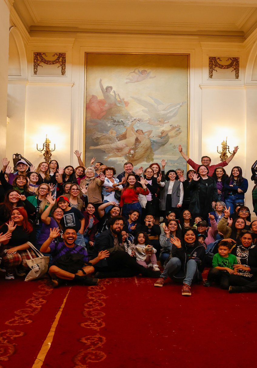 Un grupo grande de personas sonríe y saluda a la cámara en un salón con un mural.
