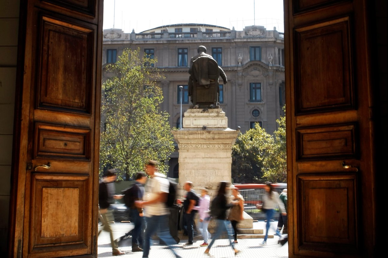 El portón de la Casa Central de la Universidad de Chile abierto hacia la Alameda, con la estatua de Andrés Bello y personas caminando.